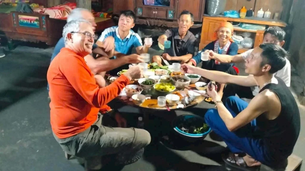 Happy travelers enjoying an authentic traditional family dinner and drinking corn liquor inside a cozy wooden Hmong homestay in Sapa.