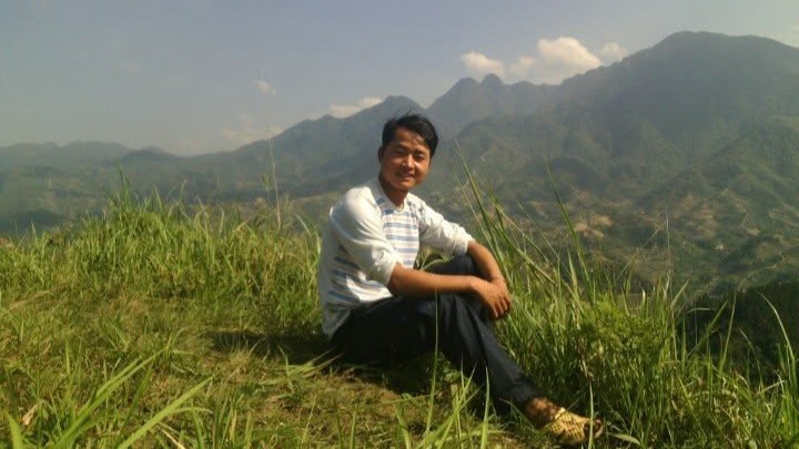 A young Thao A Sang sitting on a grassy mountain slope in Sapa wearing simple clothes and rubber sandals.